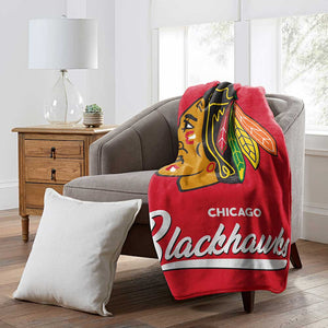 A red Chicago Blackhawks throw blanket draped over a gray armchair in a bright living room setting, with a white pillow on the floor and a wooden side table with a lamp in the background.