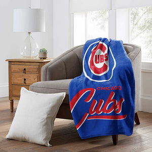 A blue Chicago Cubs throw blanket draped over a gray armchair in a bright living room, with a white pillow on the floor and a wooden side table with a lamp nearby.